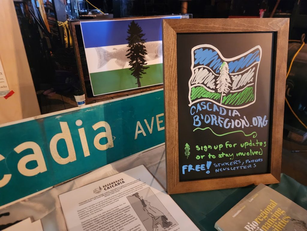 Cascadia table display with a chalkboard sign for cascadiabioregion.org, a Cascadia Doug flag print, and a Cascadia Ave street sign
